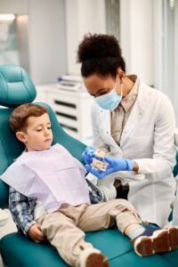 Black female dentist using denture and toothbrush during appointment with child at dentist's office.