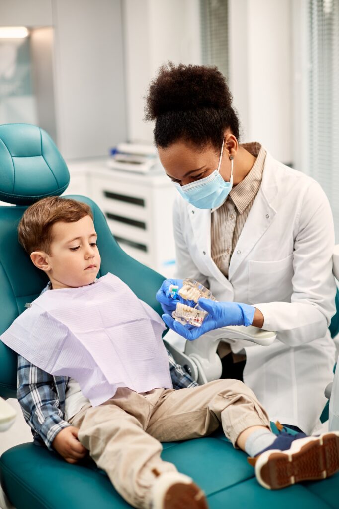 Black female dentist using denture and toothbrush during appointment with child at dentist's office.