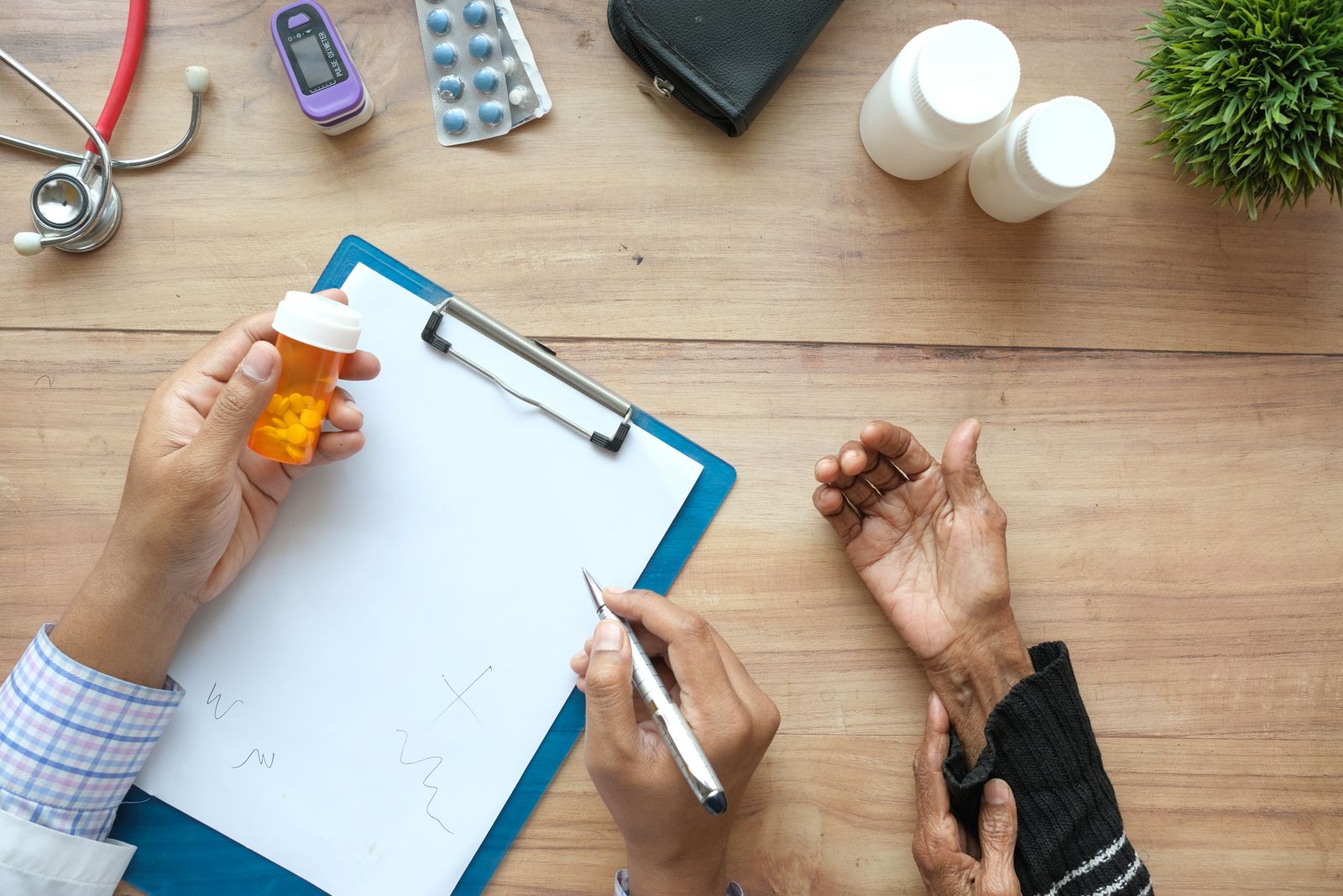 A doctor holds a pill bottle while consulting with an elderly patient. A stethoscope and prescriptions are visible