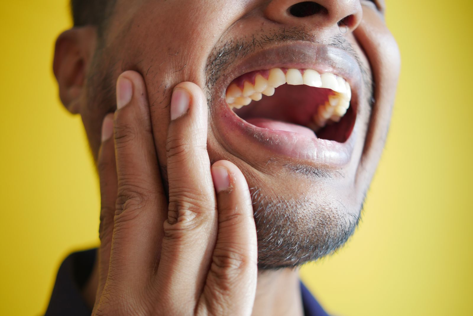 Close-up of a man holding his cheek, displaying a wide open mouth against a yellow background