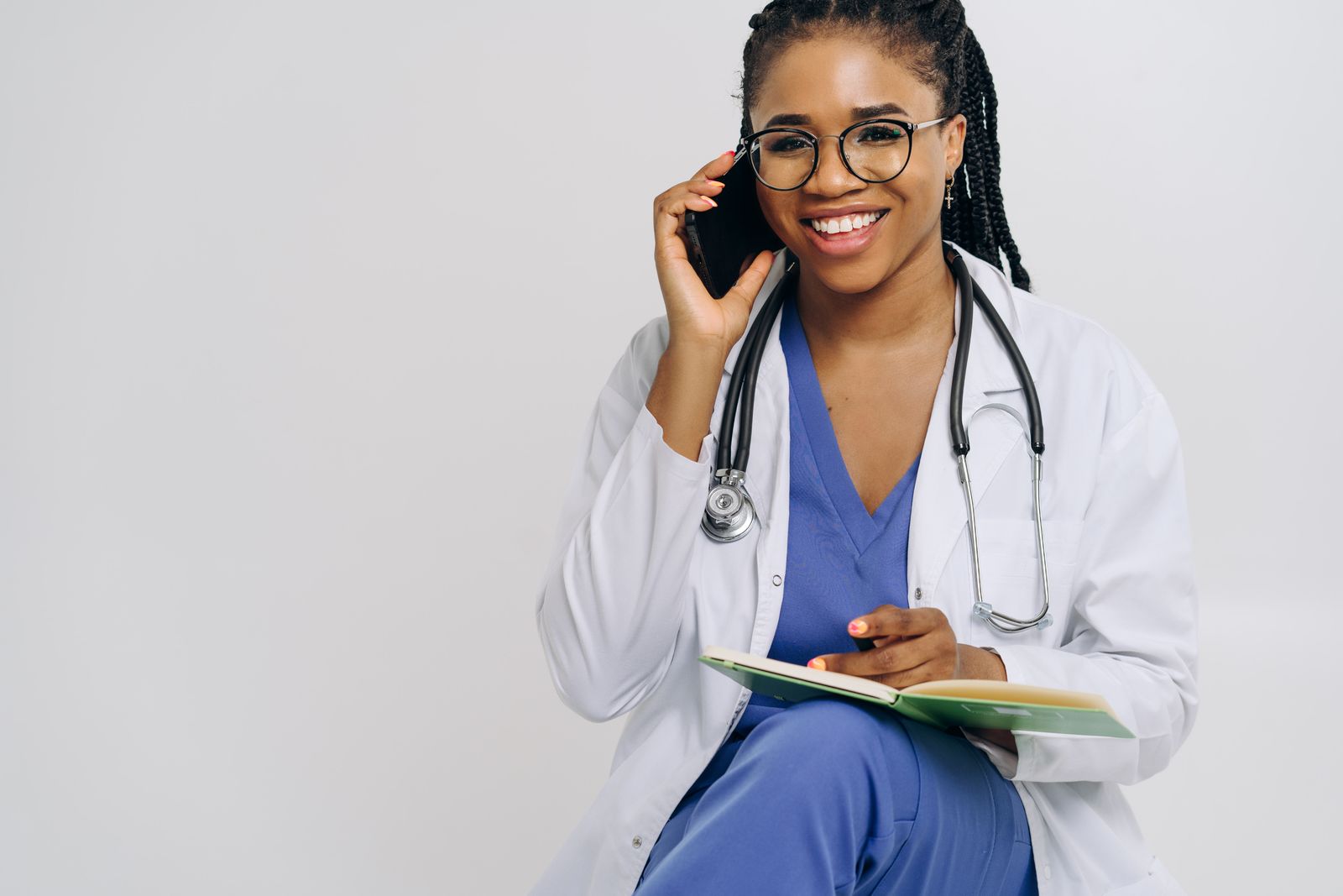 Cheerful female doctor in lab coat talking on smartphone and taking notes