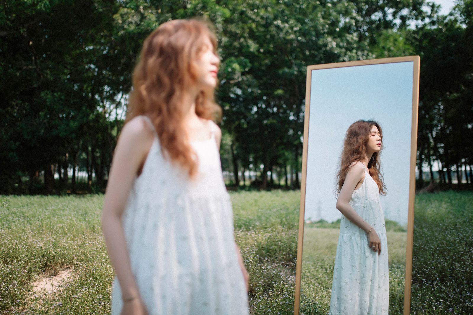 A woman in a white dress gazes at her reflection in a mirror amidst a serene outdoor setting