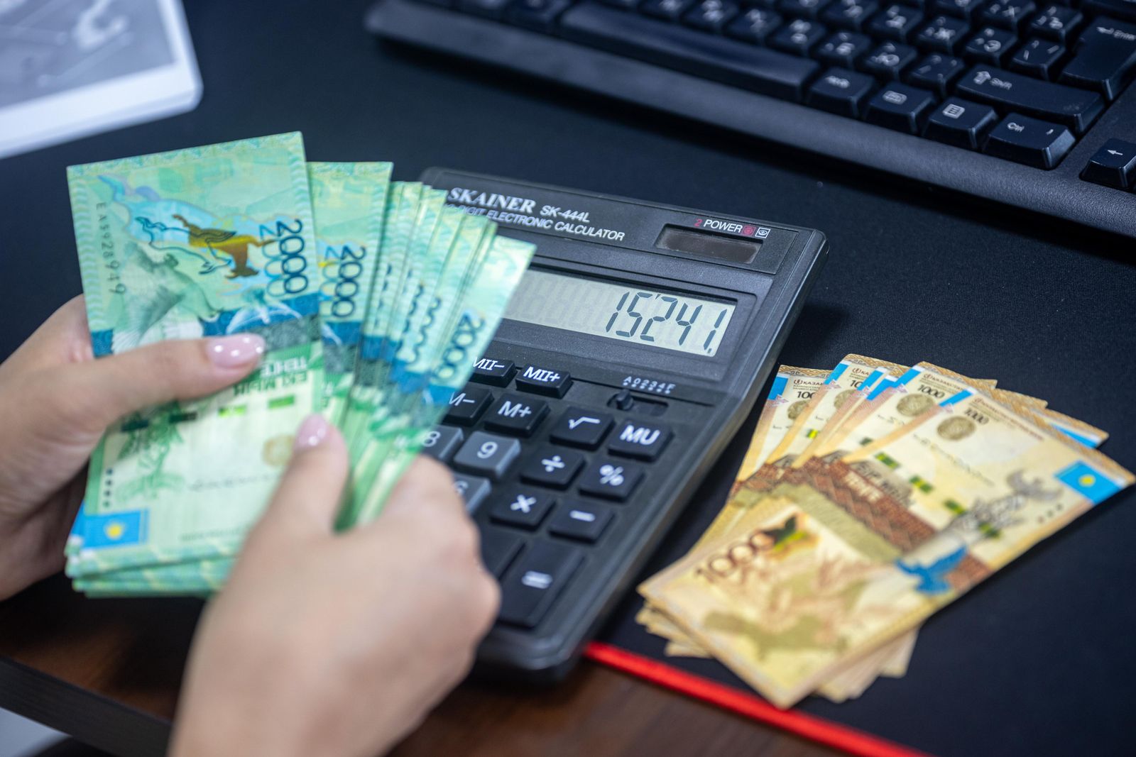 Hands counting Kazakhstani tenge while using calculator at desk