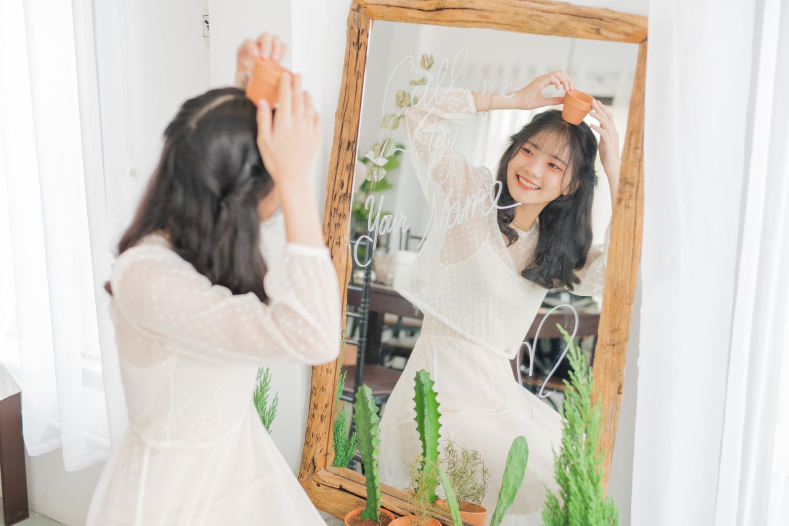 Smiling woman holding a small pot in front of mirror with plants, embracing nature indoors