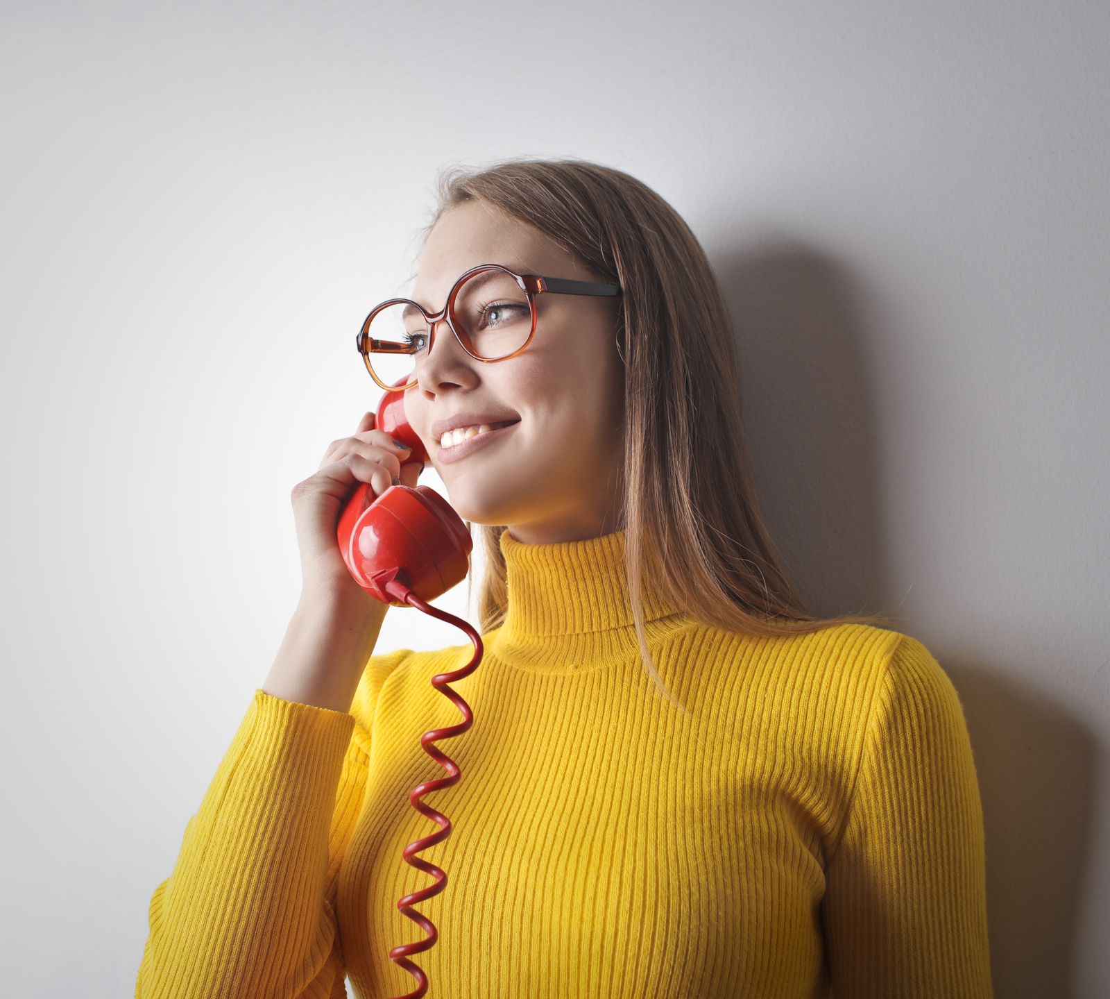 Woman in yellow sweater smiling while talking on a vintage red phone against a white wall