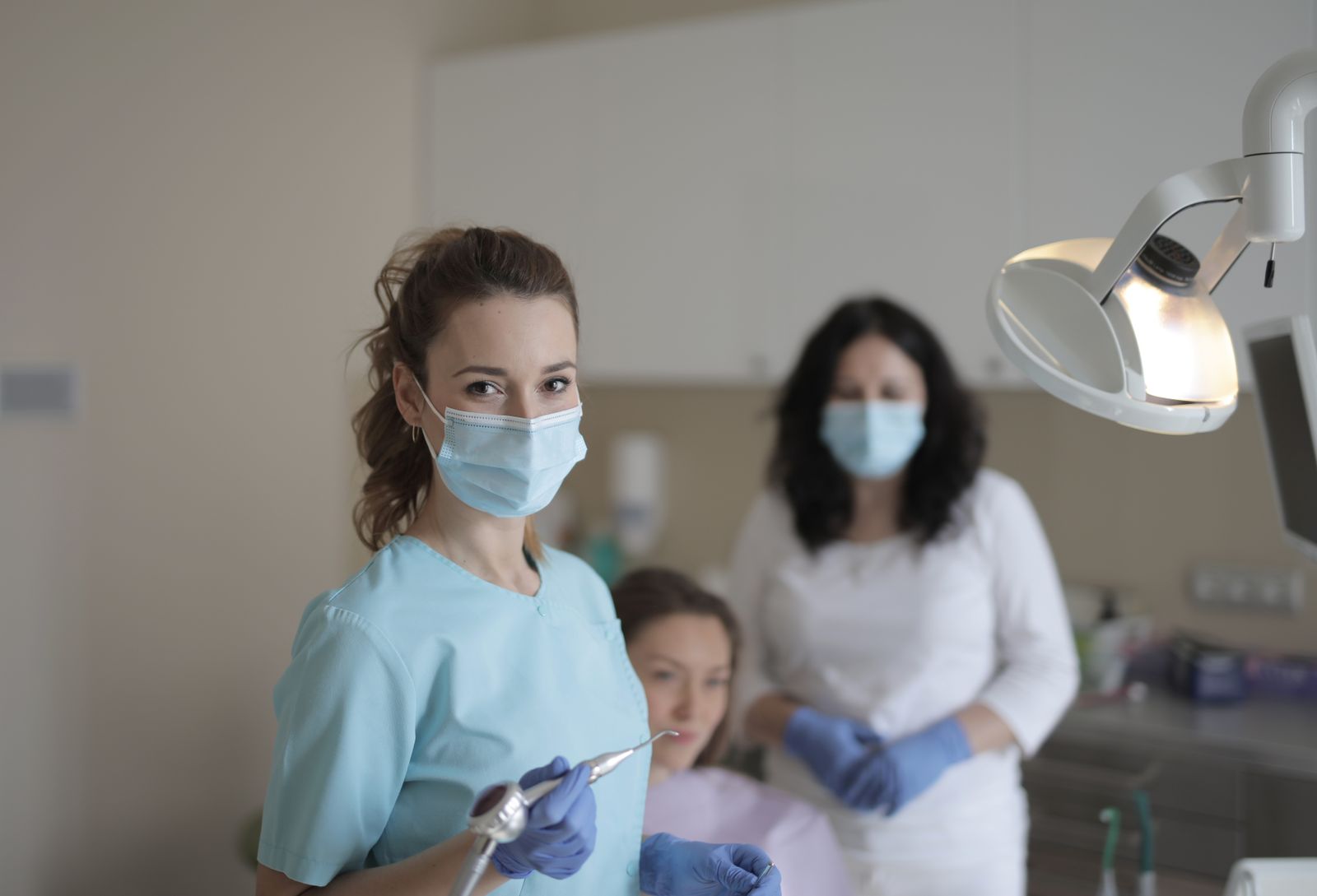Female dentists in medical masks and latex gloves wearing uniform and treating teeth of patient while standing in modern dental ro