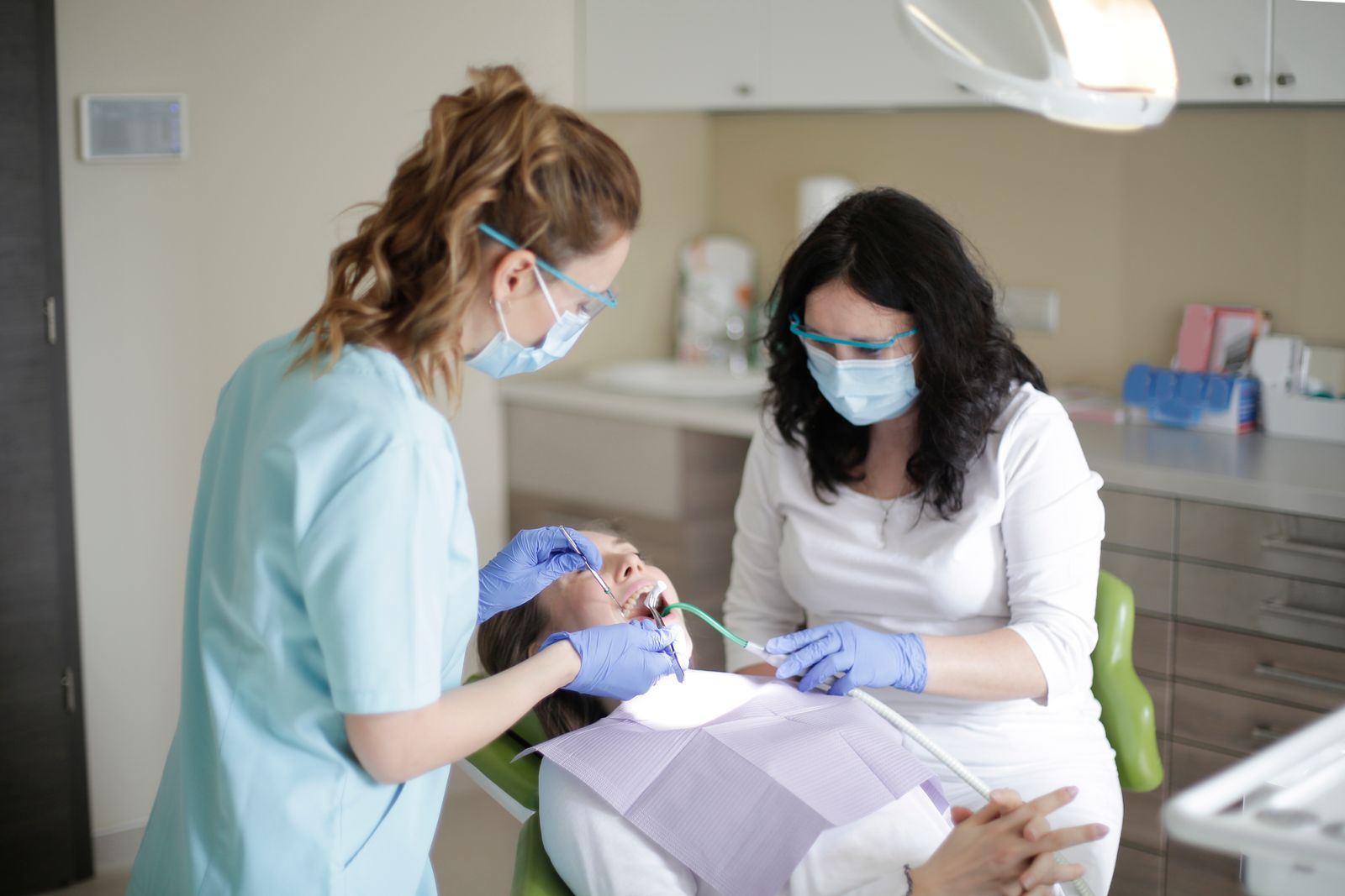Side view of female dentist in mask wearing blue uniform with latex gloves and protective glasses and assistant using dental tools