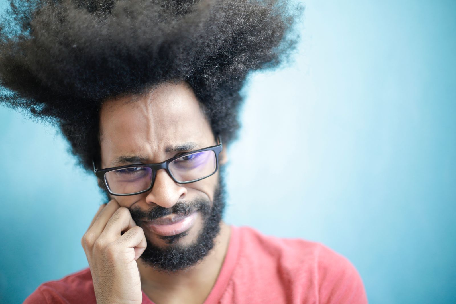 Suspicious young bearded ethnic male with creative Afro hairstyle wearing eyeglasses and pink t shirt touching cheek and looking a