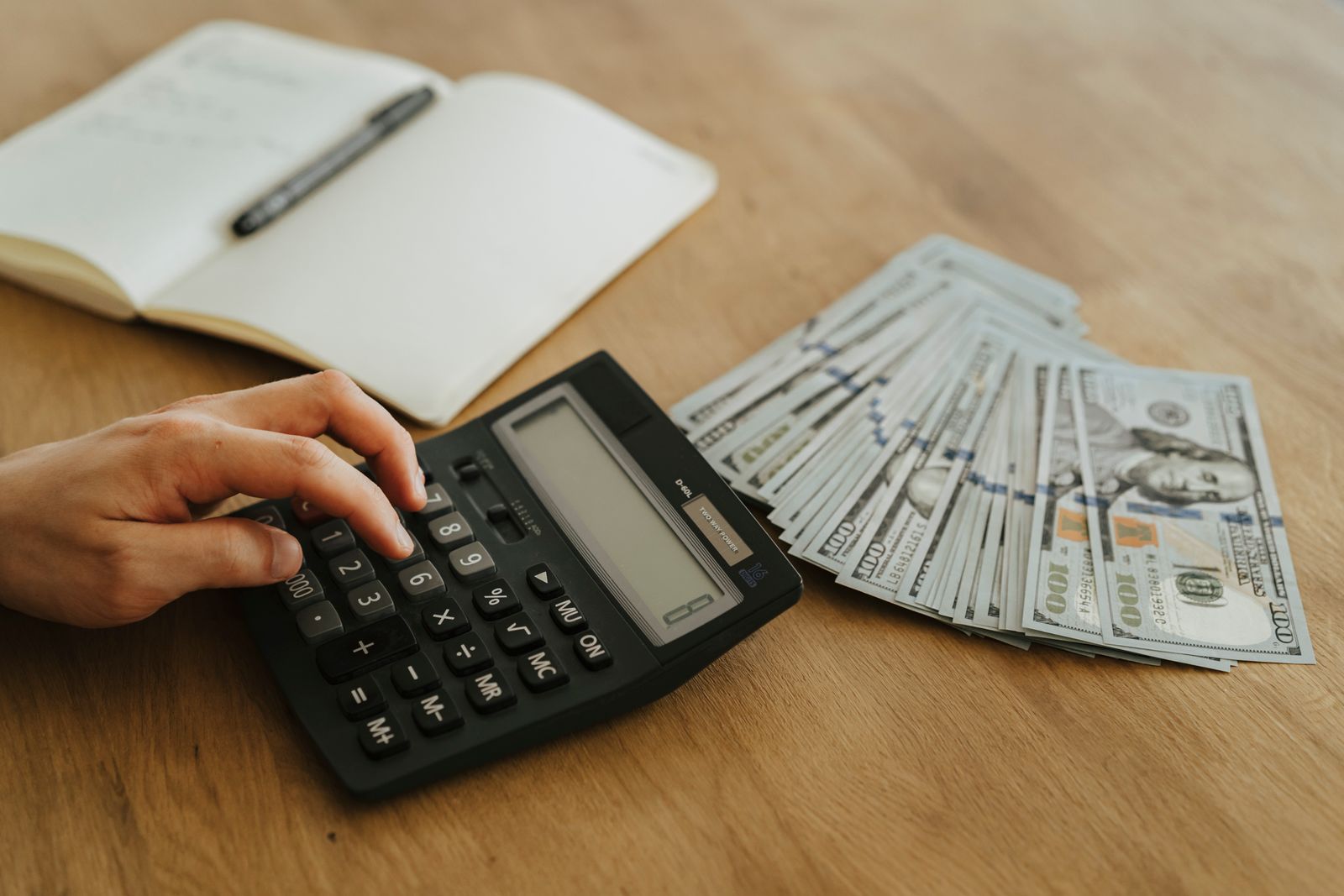 Close-up of a hand using a calculator with cash and a notebook on a wooden table