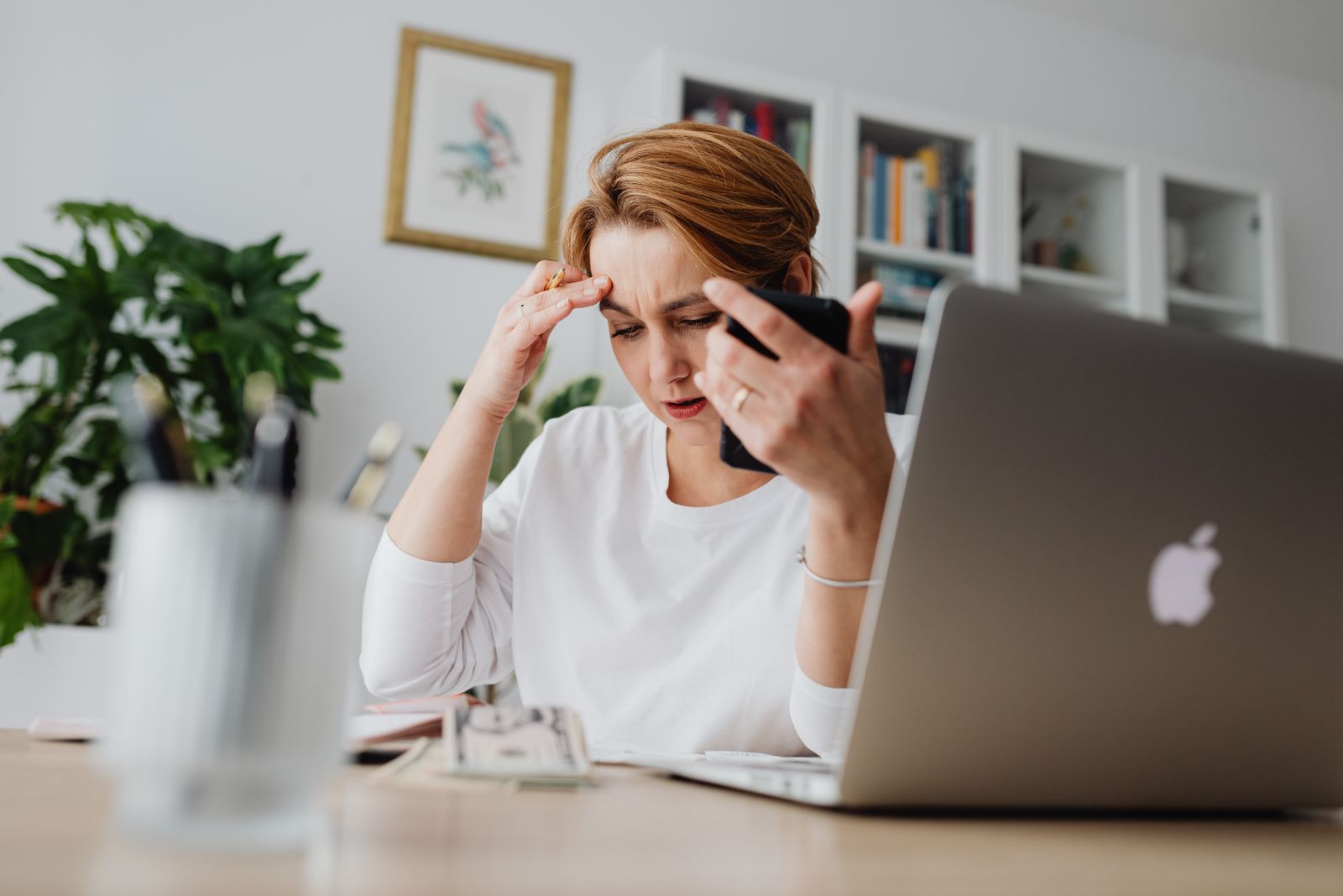 A stressed woman holds her phone, working at a desk with laptop and papers