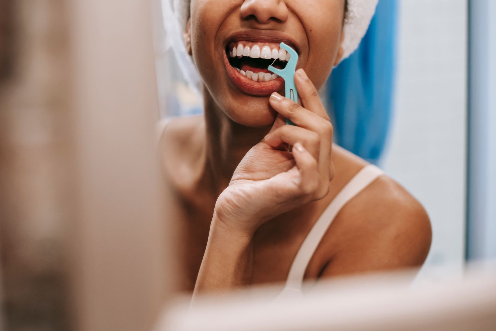 Mirror reflection of crop anonymous African American female with towel on head cleaning teeth with dental flosser in bathroom