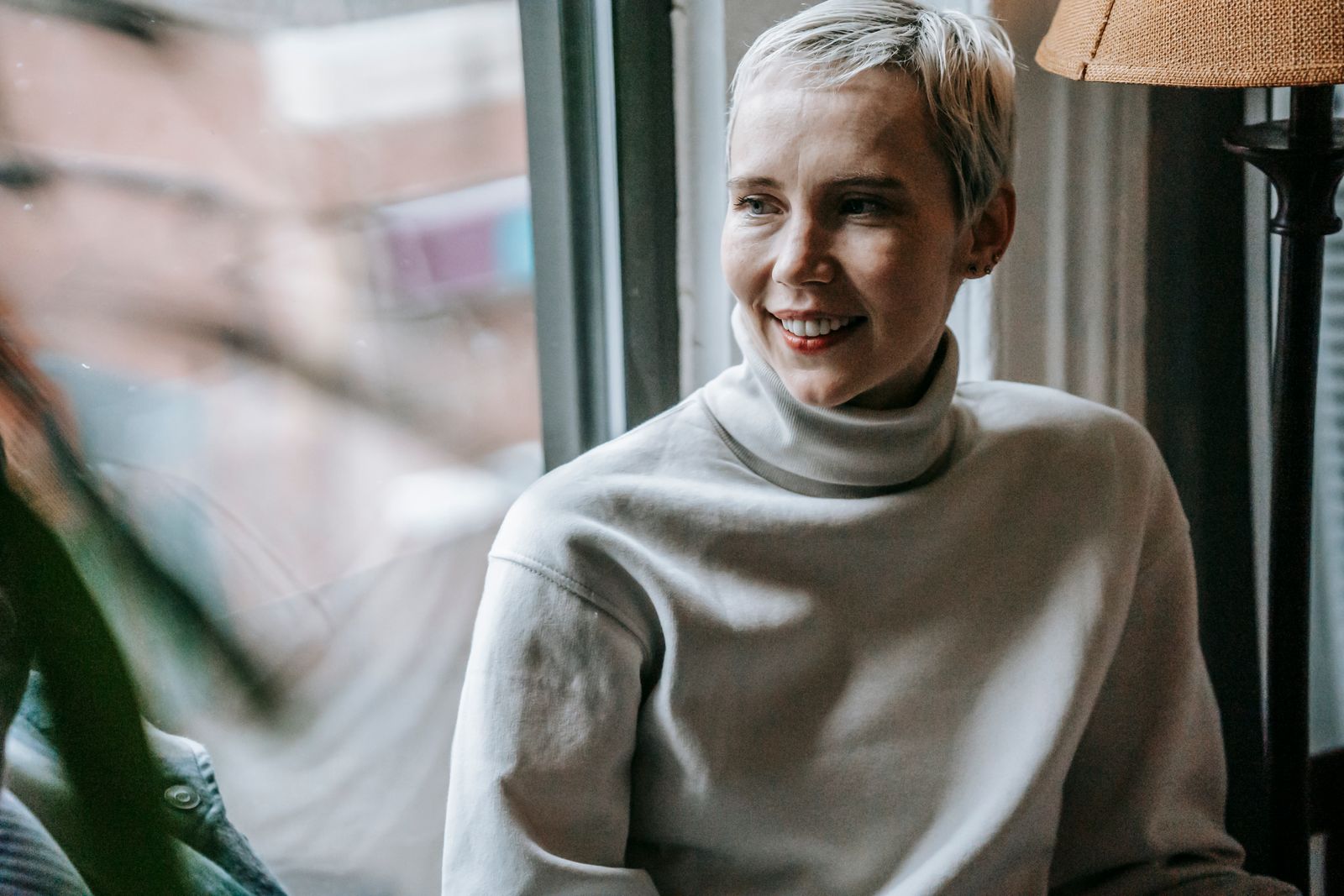 Smiling woman in casual clothes and red lipstick looking away near window and plant with green leaves near floor lamp in light roo