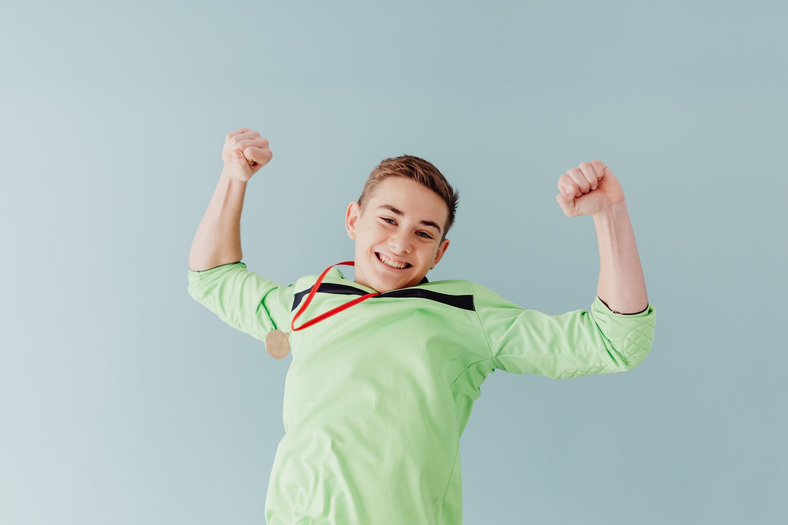 Excited teenage boy in green celebrates with a gold medal against a blue background