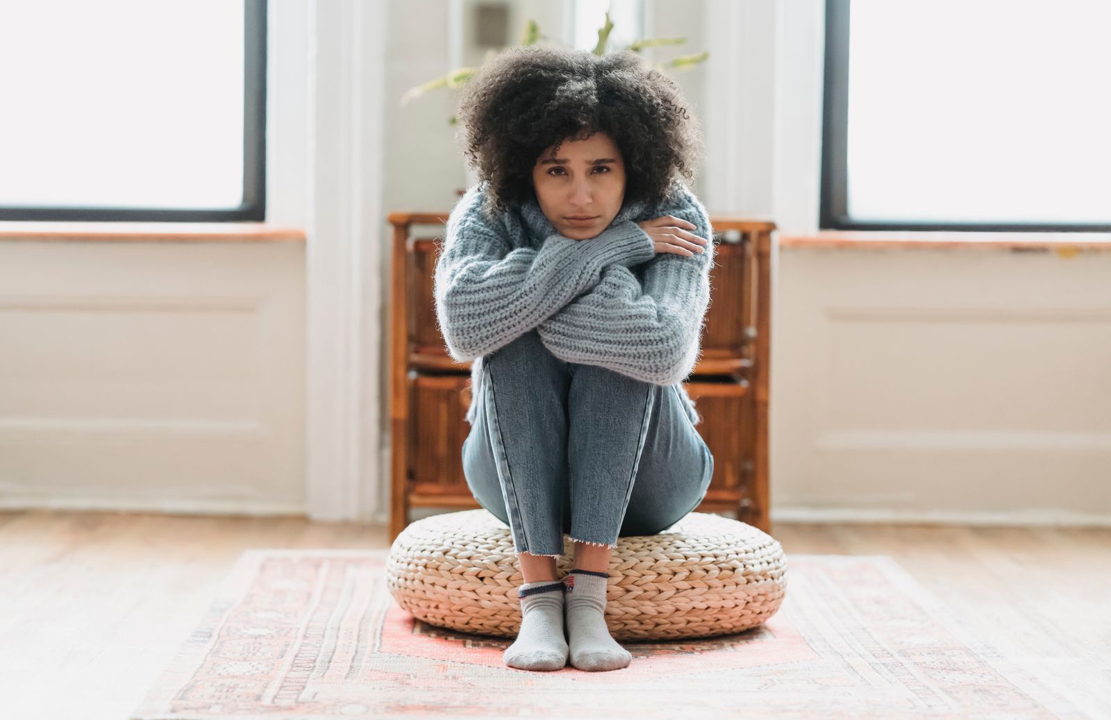 Full body of sad African American female looking at camera while sitting on rattan mat in light room at home