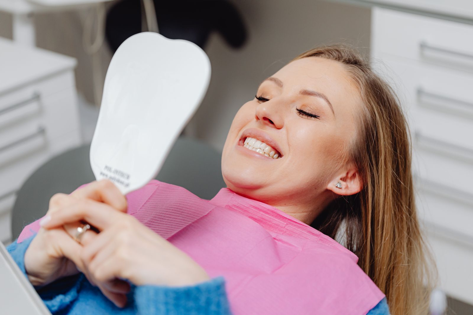 A woman at a dental clinic smiles while holding a mirror, showcasing her dental treatment results