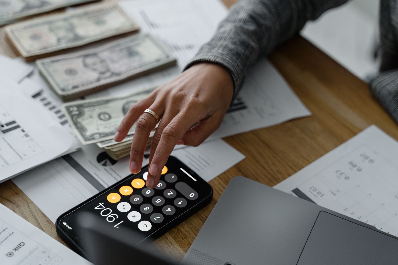 Close-up of a person using a smartphone calculator amid money and financial documents on a wooden table