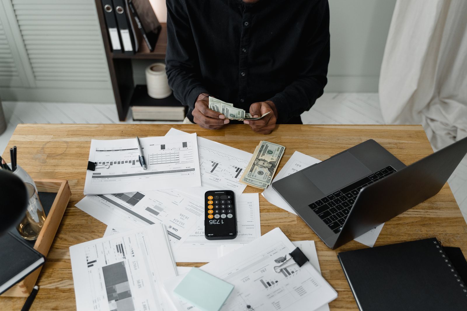 A person counts dollar bills at a desk with a laptop, papers, and calculator