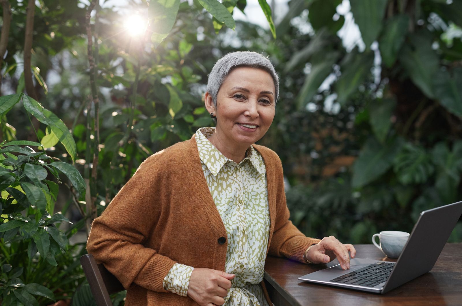 Senior woman smiling and using laptop in a lush botanical garden setting