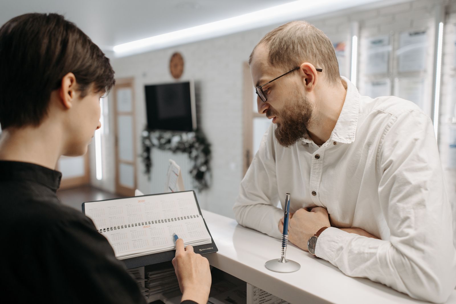 A receptionist and client converse over an appointment book at a clinic reception desk