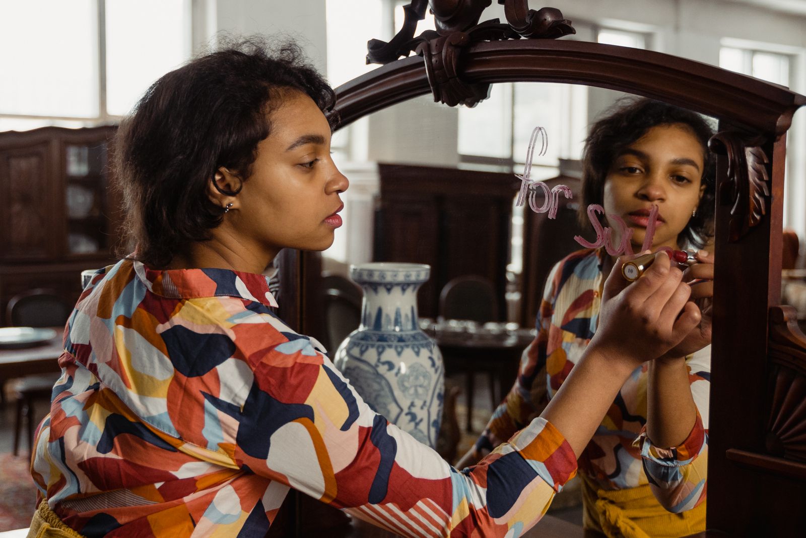 A woman writes 'for sale' on a mirror in an antique store, showcasing her reflection and vibrant printed shirt