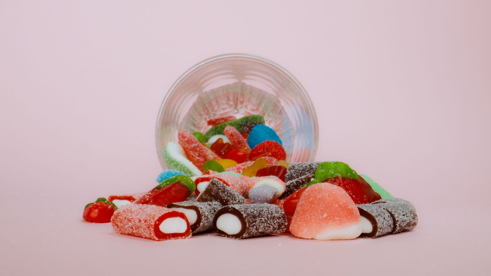 An assortment of colorful gummy candies spilling from a clear glass on a pink background