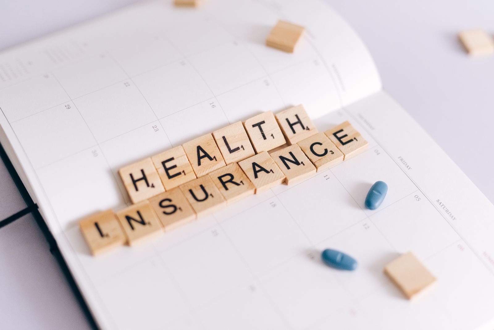 Close-up of health insurance spelled out with Scrabble tiles on a planner, alongside medication pills