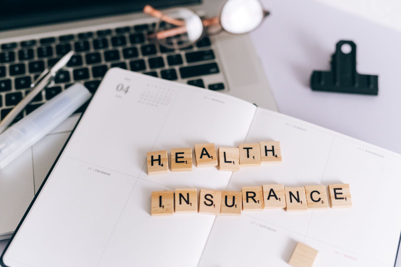 Close-up of Scrabble tiles spelling 'Health Insurance' on a planner with a laptop