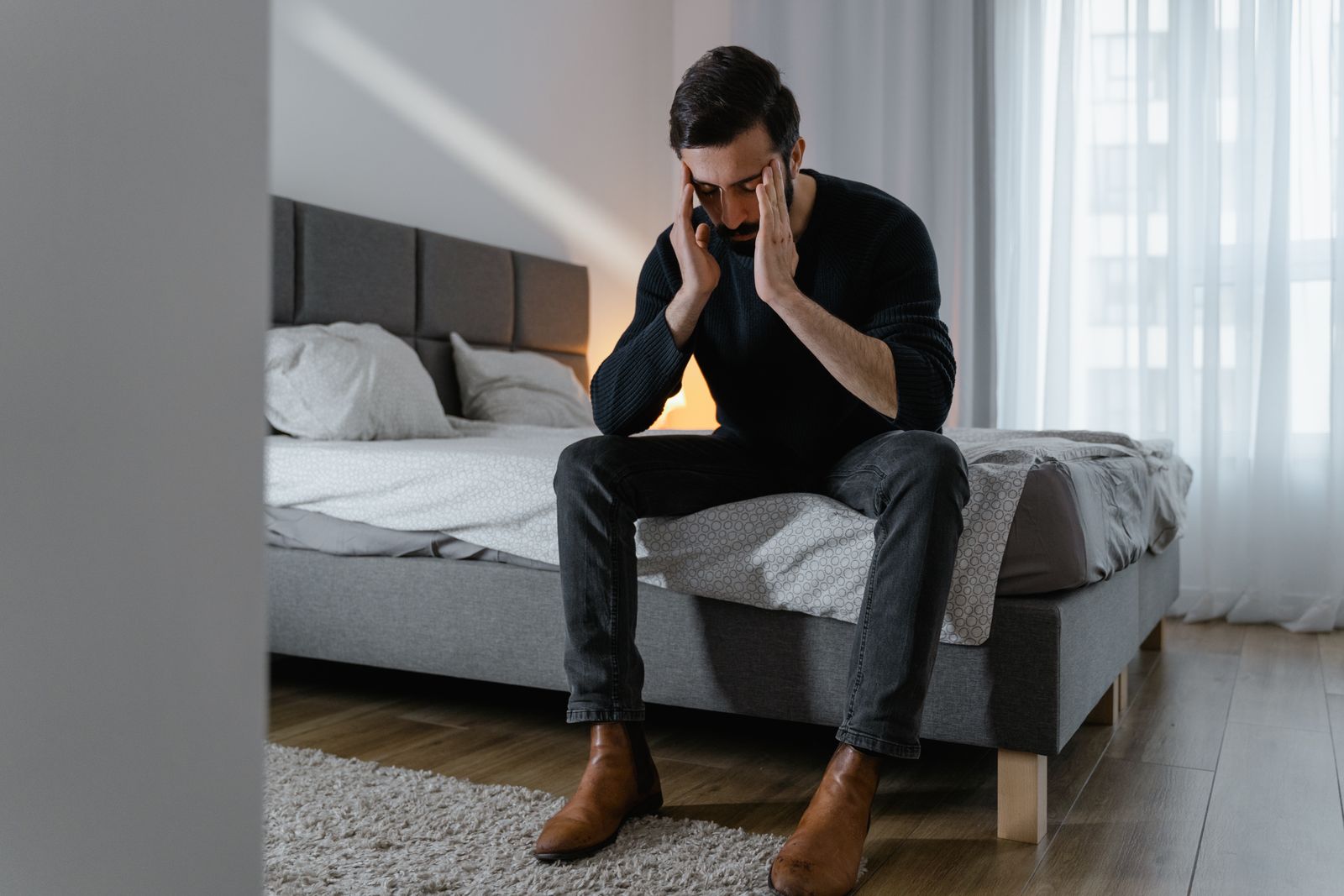 Adult man sitting on bed, appearing stressed or frustrated, holding his temples in a well-lit bedroom