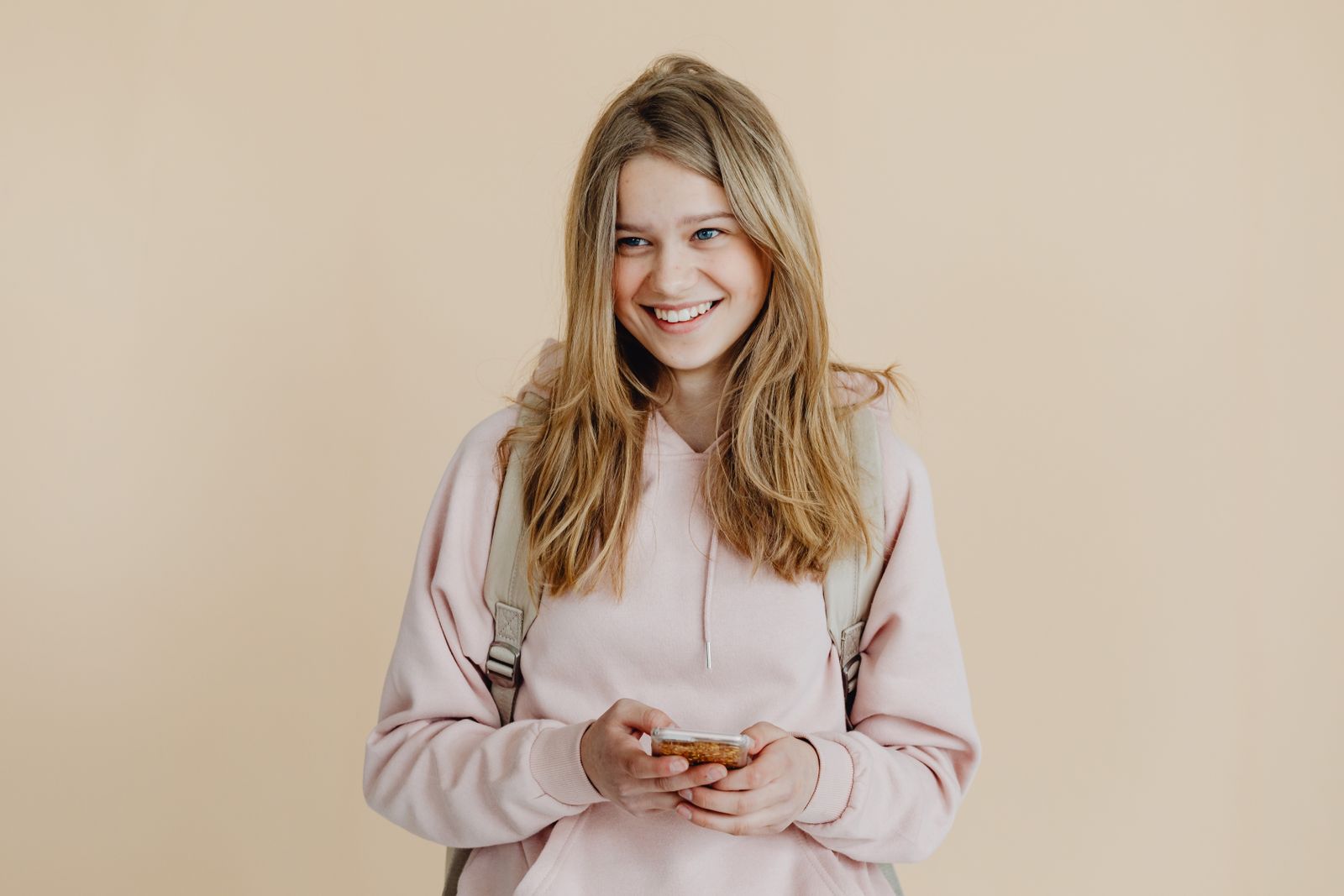 A cheerful teenager with blonde hair wearing a pink sweater and using a smartphone indoors
