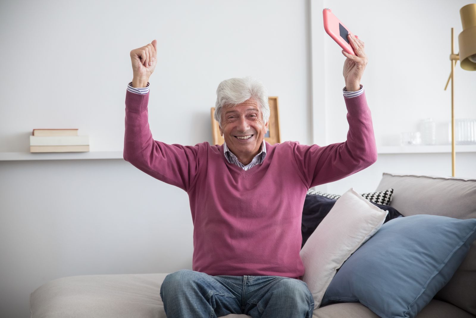 A happy senior man sitting on a sofa holding a smartphone indoors, celebrating with raised arms
