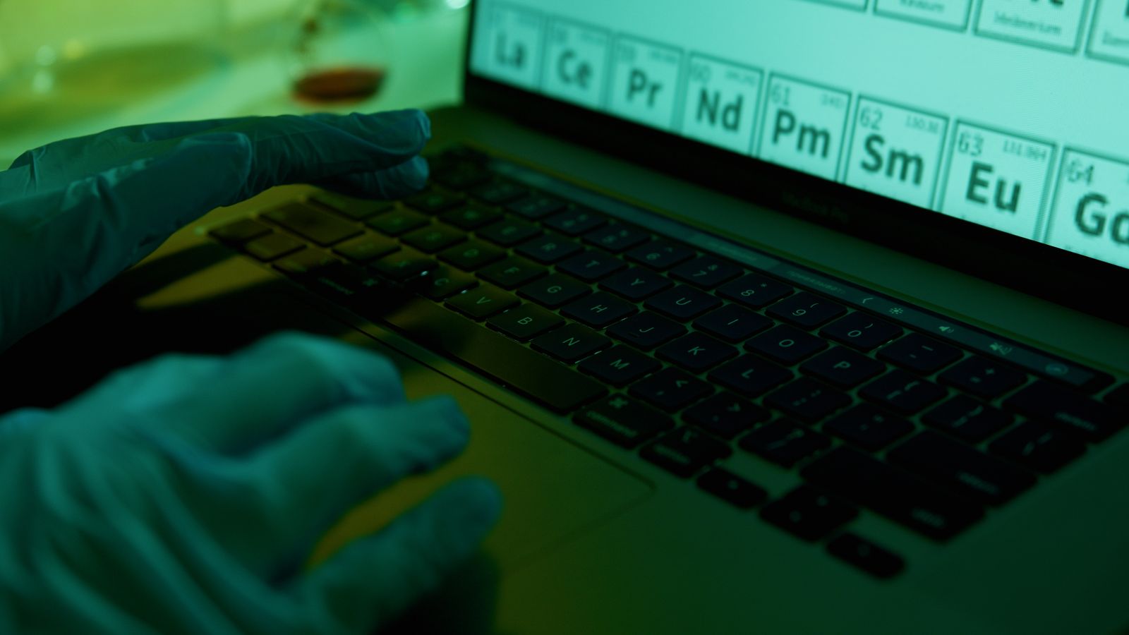 A scientist wearing gloves types data on a laptop in a laboratory setting, focused on the periodic table