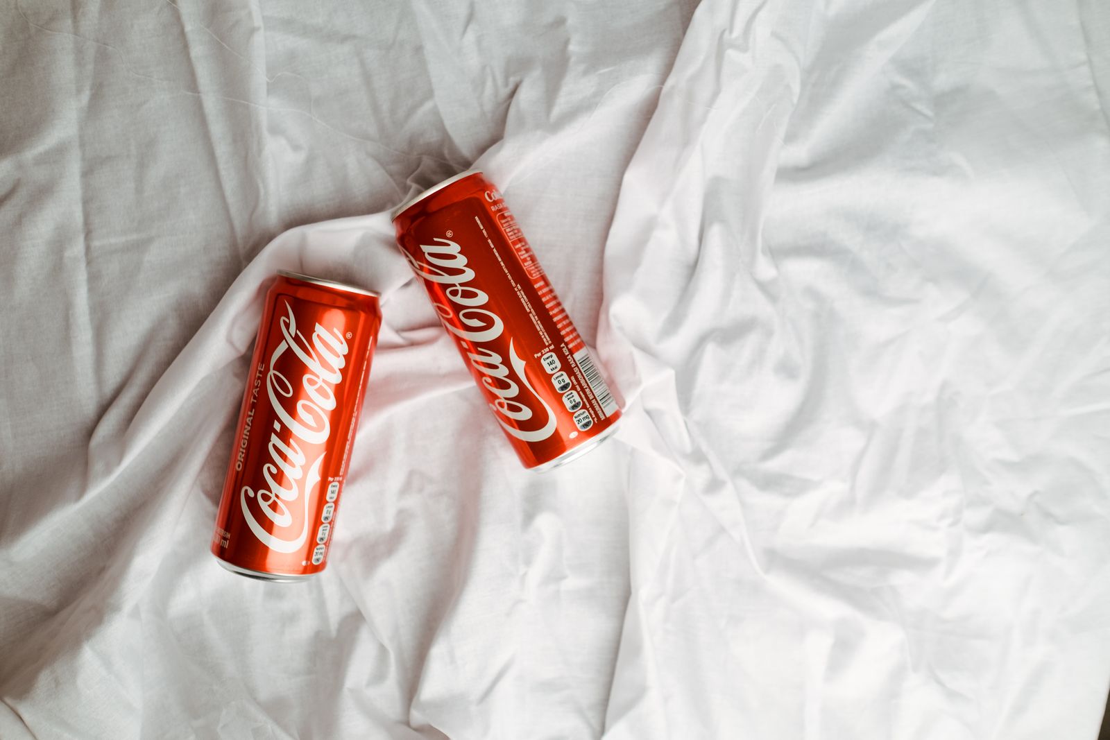 Two Coca Cola cans placed on a crumpled white textile, showcasing a classic beverage design